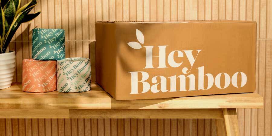 A cardboard box with the HEY BAMBOO logo, sitting on a bench next to 3 rolls of Hey Bamboo TP in colorful wrapping